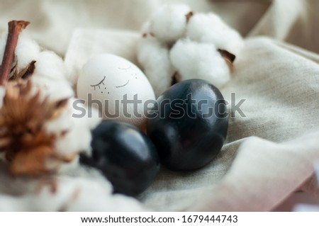 white and black Easter egg with face. Next to it is a branch of cotton on a gray linen background