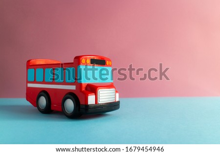 A red toy bus, with black wheels and blue windows. The bus is isolated on its own, against a plain pink background. There are slots for toy people to go on top of the bus.