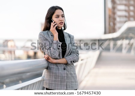 Stock photo of a young business woman having a conversation on the phone. She is serious. She is standing on the street. She is wearing casual clothing.