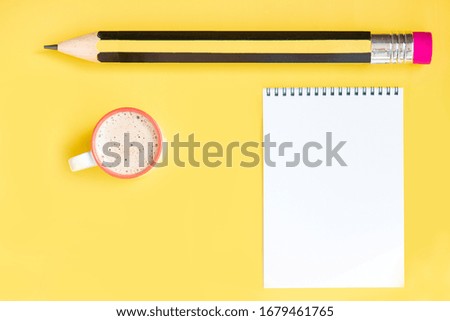 Cup of cappuccino, paper notepad and huge pencil on the yellow background.