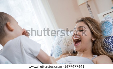 Beutiful young mother and toddler son enjoying each other in the morning. Smiling, laughing and playing in bed with child. Family happiness concept. Bright, sunny warm day.