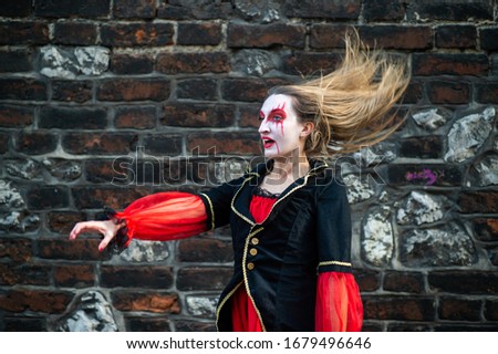 Quaint female in witch costume with an ominous expression of his face is engaged in magic and conjures putting her hands forward against the background of the brick wall