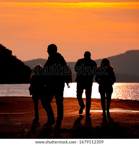 Silhouette of family walking along the seashore on orange sunset, four adults and one child, golden path on water surface, cliffs and mountain silhouettes on background