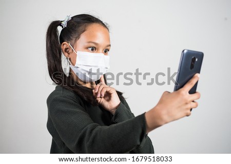Girl with a prevention mask is entertained at home taking photos with the phone with facial gestures. Concept of home quarantine during the global infection.