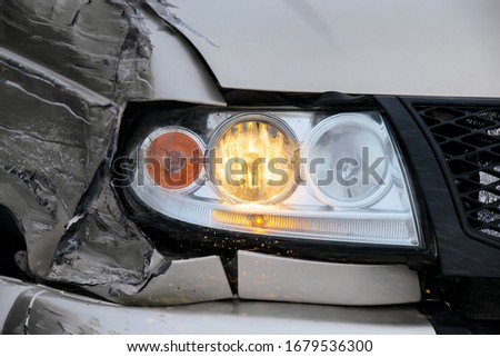 A closeup of broken headlights on the front of a car in an accident. The headlights continue to glow. The left side of the car is badly damaged. Road safety theme.