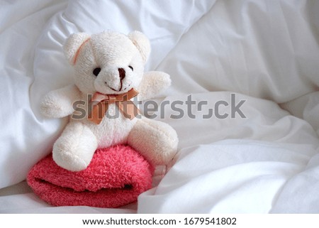 A soft toy bear, white in color, sits on a pink pillow.  White linen.