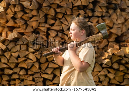 Portrait of handsome caucasian girl in yellow t-shirt and black trousers with an axe against the background of a large pile of firewood, chop wood in the village