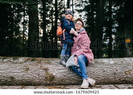 picture of lovely young caucasian female with pretty face, short dark hair, big eyes sits on the felled trees with her child in a forest