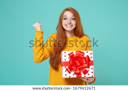 Smiling redhead girl in yellow sweater posing isolated on blue turquoise background. Valentine's Day Women's Day birthday concept. Hold white red present box with gift ribbon bow doing winner gesture