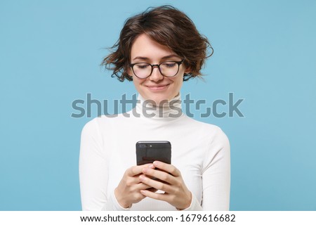 Smiling young brunette woman girl in casual white clothes, eyeglasses posing isolated on pastel blue background. People lifestyle concept. Mock up copy space. Using mobile phone, typing sms message Royalty-Free Stock Photo #1679616682