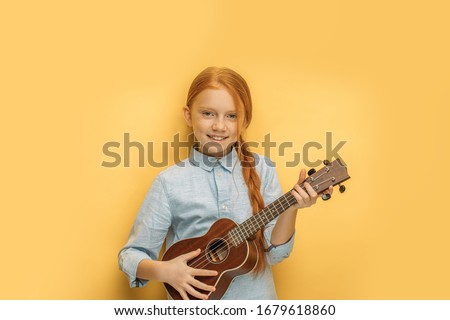 beautiful caucasian girl learns to play ukulele isolated over yellow background. attractive child with natural red hair hold small guitar, ukulele in hands and look at camera, smile, enjoy music Royalty-Free Stock Photo #1679618860