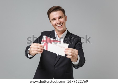 Cheerful young business man in classic black suit shirt posing isolated on grey wall background studio portrait. Achievement career wealth business concept. Mock up copy space. Hold gift certificate