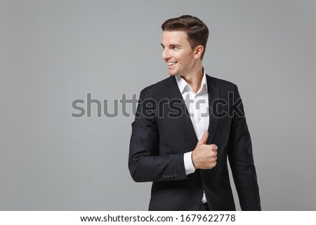 Smiling young business man in classic black suit shirt posing isolated on grey background in studio. Achievement career wealth business concept. Mock up copy space. Showing thumb up looking aside