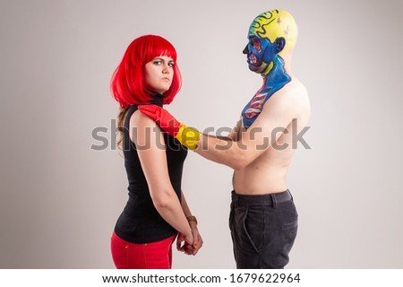 A strange man with makeup on his head and shoulder in red-yellow gloves, black pants and boots, no T-shirt with young caucasian female with re hair, red trousers, black t-shirt