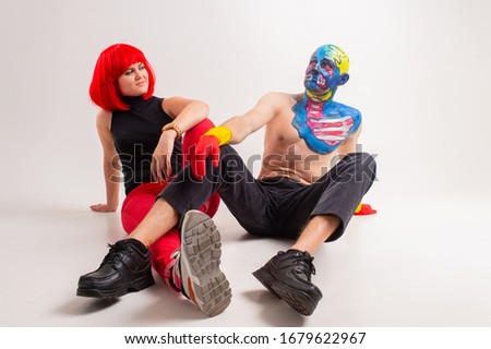 A strange man with makeup on his head and shoulder in red-yellow gloves, black pants and boots sits on a floor with young caucasian female with red hair, red trousers, black t-shirt