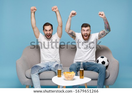 Screaming guys friends in white t-shirt sit on couch isolated on blue background. Sport leisure concept. Cheer up support favorite team expressive gesticulating with rising hands doing winner gesture