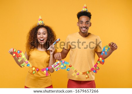 Excited young friends couple african american guy girl in casual clothes birthday hat isolated on yellow orange background. Holiday party lifestyle concept. Celebrating holding pipe, paper garland
