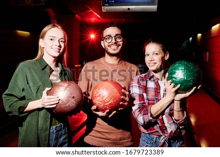 Three young intercultural friendly bowling players holding balls while standing in dark room on background of alleys