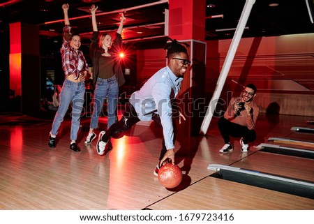 Cheerful active guy of African ethnicity going to throw bowling ball while two happy girls raising arms and young man photographing him