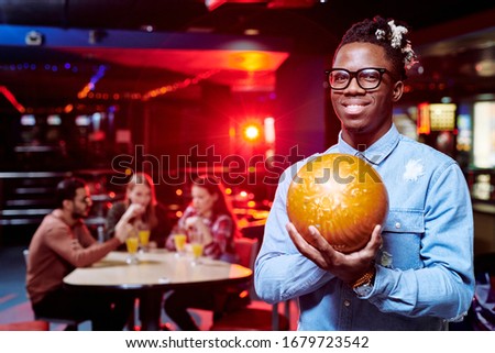 Happy young African man with bowling ball standing in front of camera during play on background of his friends having drinks