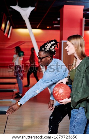 African guy with stretched arm explaining his girlfriend how to throw bowling ball on track or alley while playing at leisure center