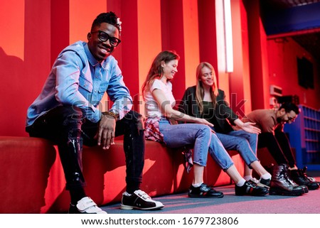 Cheerful young African man in casualwear and bowling shoes looking at you while sitting on background of his happy friends