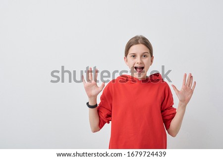 Friendly and cheerful teenage girl in red sweatshirt looking at you with her palms open and expressing gladness over white background