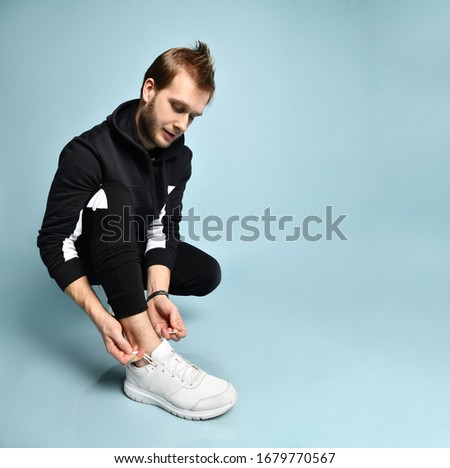 Handsome hipster guy with cool hairstyle, in black tracksuit, bracelet and white sneakers. He squatting and tying shoelaces, posing against blue studio background. Trendy outfit. Close up, copy space
