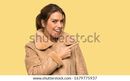 Blonde woman with a coat pointing to the side to present a product over isolated yellow background