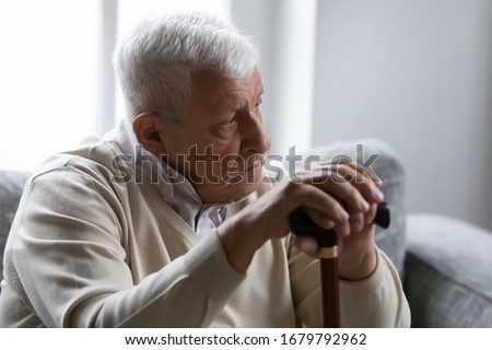 Serious gray-haired man hold wooden walking stick sit on couch in living room alone look at distance feels upset and lonely. Nursing home for older disabled people, senile diseases, healthcare concept Royalty-Free Stock Photo #1679792962