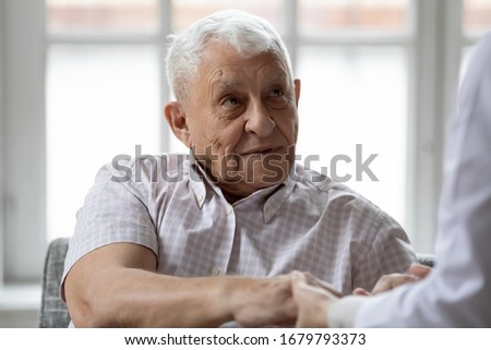 Woman nurse hold hand of elderly grey-haired man showing care and bond close up, doctor in white coat talk with aged patient sitting together indoor, concept of support, caregiving and nursing service Royalty-Free Stock Photo #1679793373