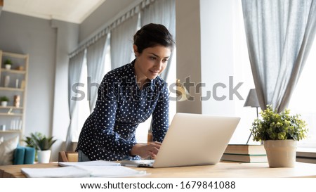 Smiling Indian girl standing at desk, working on laptop, looking at screen, browsing, searching information in internet, young female student studying online at home, chatting in social network