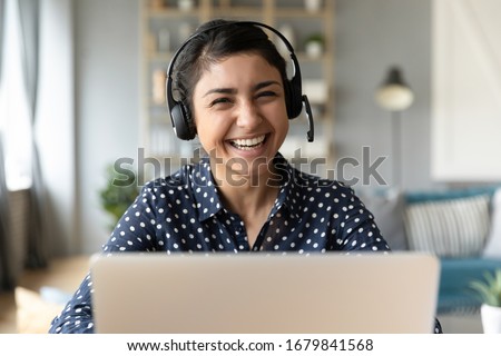Head shot portrait smiling Indian woman wearing headphones posing for photo at workplace, happy excited female wearing headset looking at camera, sitting at desk with laptop, making video call Royalty-Free Stock Photo #1679841568