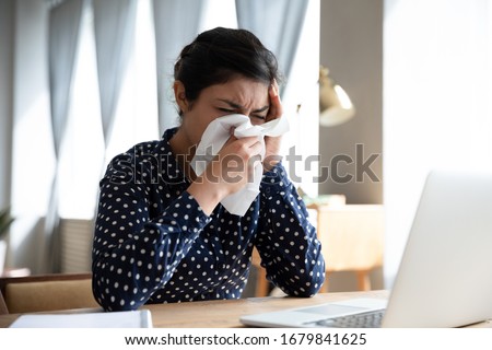 Unhealthy Indian woman blowing running nose, sitting at workplace, exhausted businesswoman or student feeling bad, suffering from fever, holding handkerchief, allergic reaction or seasonal infection Royalty-Free Stock Photo #1679841625