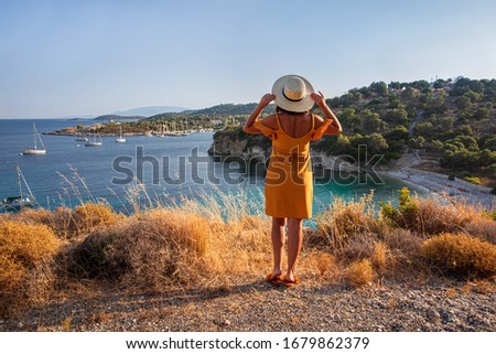 Kastos island happy young woman in a yellow dress and a straw hat enjoying scenic view of the amazing summer seascape with calm sea, anchored sailboats, beautiful nature - Ionian sea, Greece.