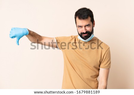 Caucasian man with beard protecting from the coronavirus with a mask and gloves over isolated background showing thumb down with negative expression