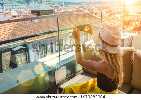 Young traveler woman with smartphone sitting on roof cafe in Lisbon and takes pictures of city under sunset sky.
