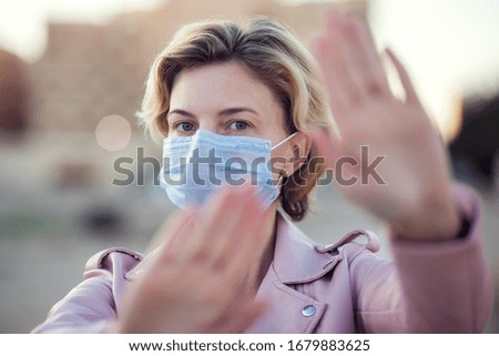 A portrait of woman with short blond hair with medical face mask showing stop sign with hands outdoor. People, healthcare and medicine concept