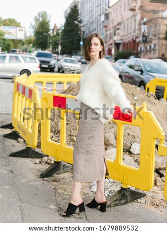 Woman in beige sweater and skirt staying in front of road repair sign. Street Fashion