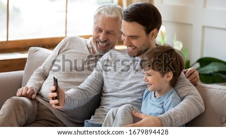 Smiling three generations of men sit on couch in living room make self-portrait picture on smartphone, happy little boy with young dad and mature grandfather take selfie on cellphone gadget together Royalty-Free Stock Photo #1679894428