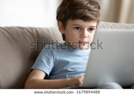 Concentrated smart little preschooler boy sit on sofa at home browsing wireless Internet on laptop, small serious child watch video use modern computer gadget, kids and technology, education concept