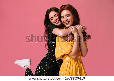 Curly girls smiling and hugging on pink background. Beautiful girlfriends with fashionable make-up in light dresses smile on isolated