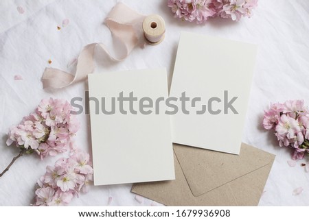 Wedding stationery mock-up scene. Blank greeting cards, envelope on linen tablecloth background with pink blossoming cherry tree branches and ribbon. Feminine still life composition. Flat lay,top view Royalty-Free Stock Photo #1679936908