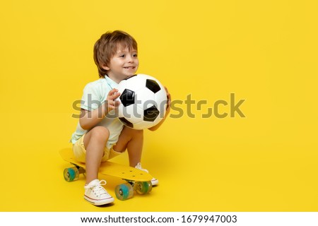 Amazed little boy play with soccer ball while sitting on pennyboard or skateboard over yellow background.
