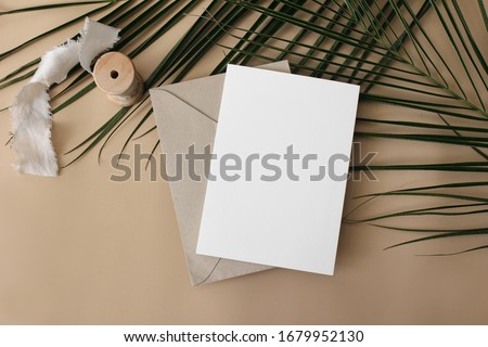 Summer wedding stationery mock-up, desk scene. Blank greeting card, envelope, ribbon and green palm leaves on beige table background. Tropical exotic styled photo, web banner. Flat lay, top view.