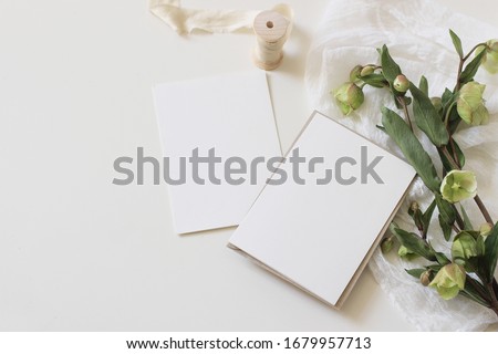 Wedding spring styled stock photo. Feminine desktop mockup scene with green hellebores flowers, silk ribbon, muslin cloth and blank paper greeting cards on white table background. Flat lay, top view.