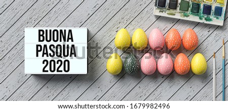 easter eggs with message HAPPY EASTER 2020 in Italian and lightbox with number 2020 surrounded by brushes and water color boxes on wooden background