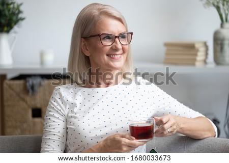 Head shot close up portrait happy stylish older lady in eyewear relaxing alone with cup of tea on comfortable sofa at home. Smiling middle aged woman enjoying weekend leisure free time, dreaming.