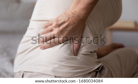 Close up older retired woman sitting on bed, touching lower back, suffer from painful feelings. Unhealthy middle aged lady having lumbar discomfort, pain in muscles after waking up, rear view. Royalty-Free Stock Photo #1680034084