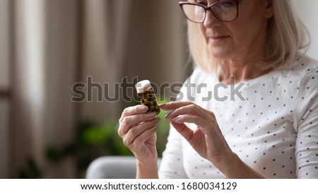 Close up concentrated middle aged woman wearing eyeglasses, holding bottle with pills in hands, reading instruction to medicine. Focused mature lady examining drug prescription label at home. Royalty-Free Stock Photo #1680034129